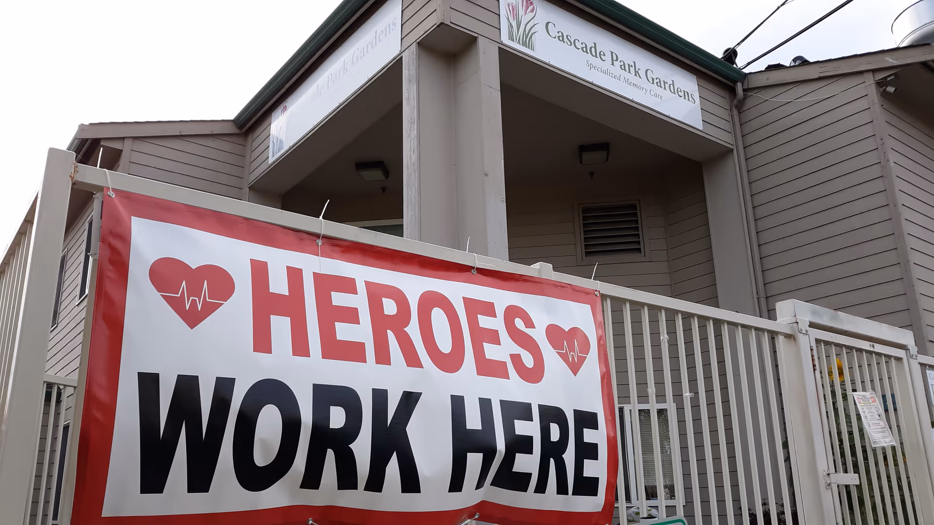A large 'HEROES WORK HERE' banner hangs on a fence in front of the Cascade Park Gardens building entrance.