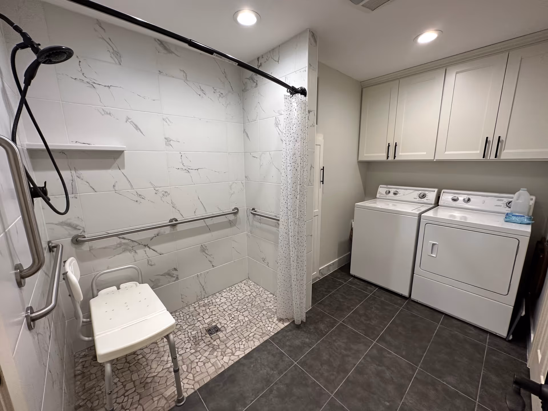 A combined laundry and accessible shower room featuring a walk-in shower with a white shower chair, grab bars, and a handheld showerhead. Adjacent to the shower are a white washing machine and dryer beneath white cabinets. The floor is tiled in dark gray, and the shower walls have white tiles with gray marble-like patterns.
