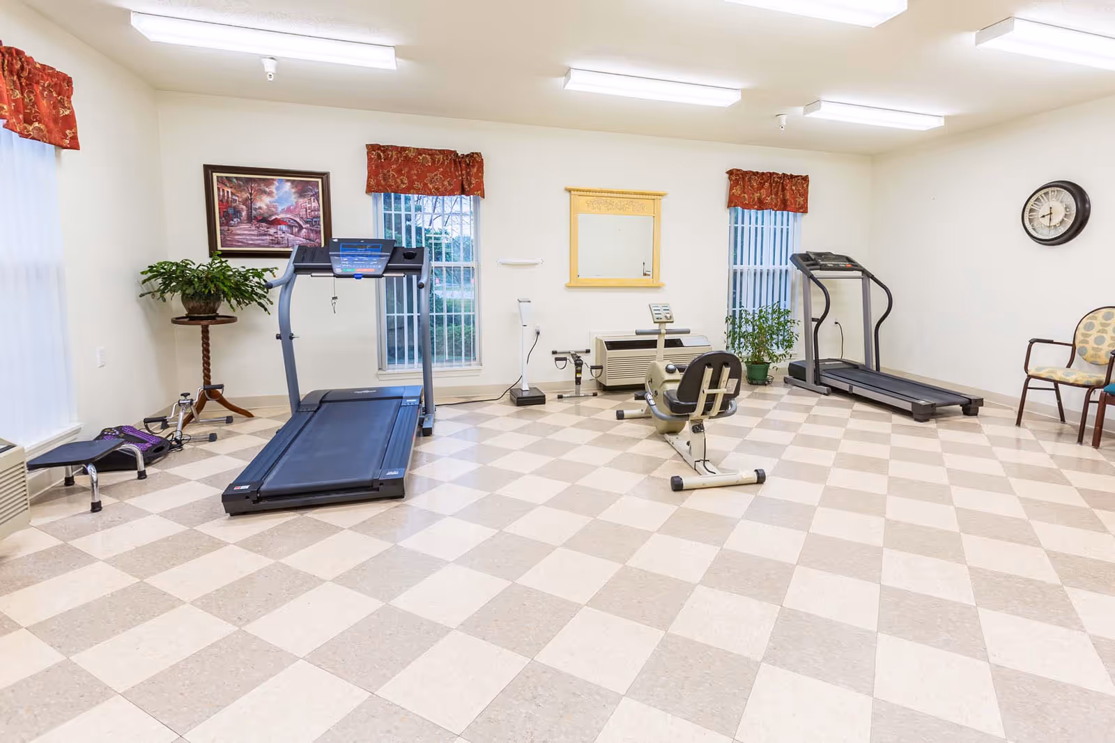 Well-lit exercise room with two treadmills, a recumbent bike, and chairs on a checkered tile floor.