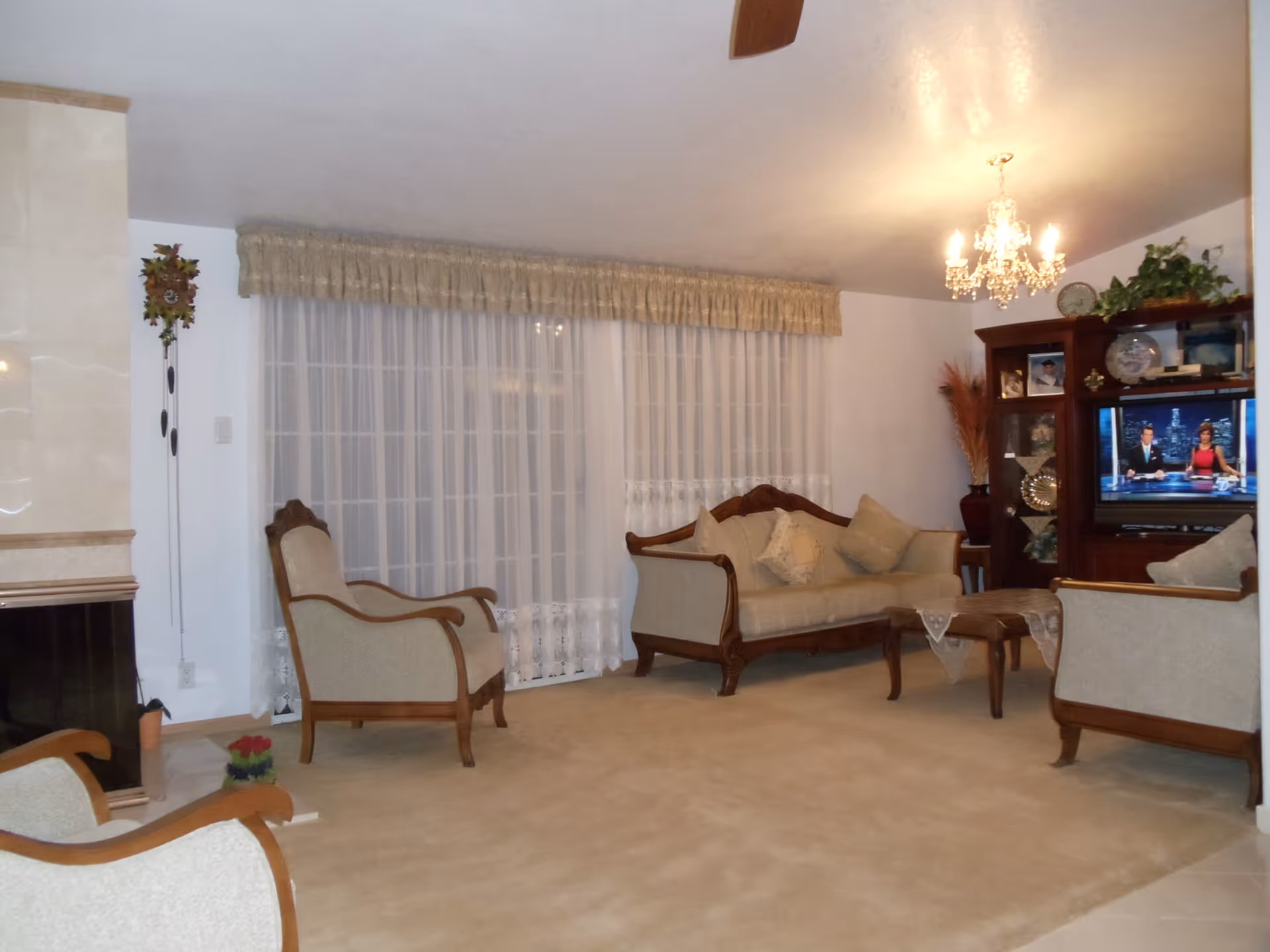 A cozy living room with beige carpet and classic wooden furniture including a sofa and armchairs with beige cushions. A wooden coffee table with lace doilies is in the center. A chandelier hangs from the ceiling, illuminating the room. Sheer white curtains cover large windows, and a wooden entertainment center holds a television displaying a news broadcast. Decorative items and plants are placed on the shelves.