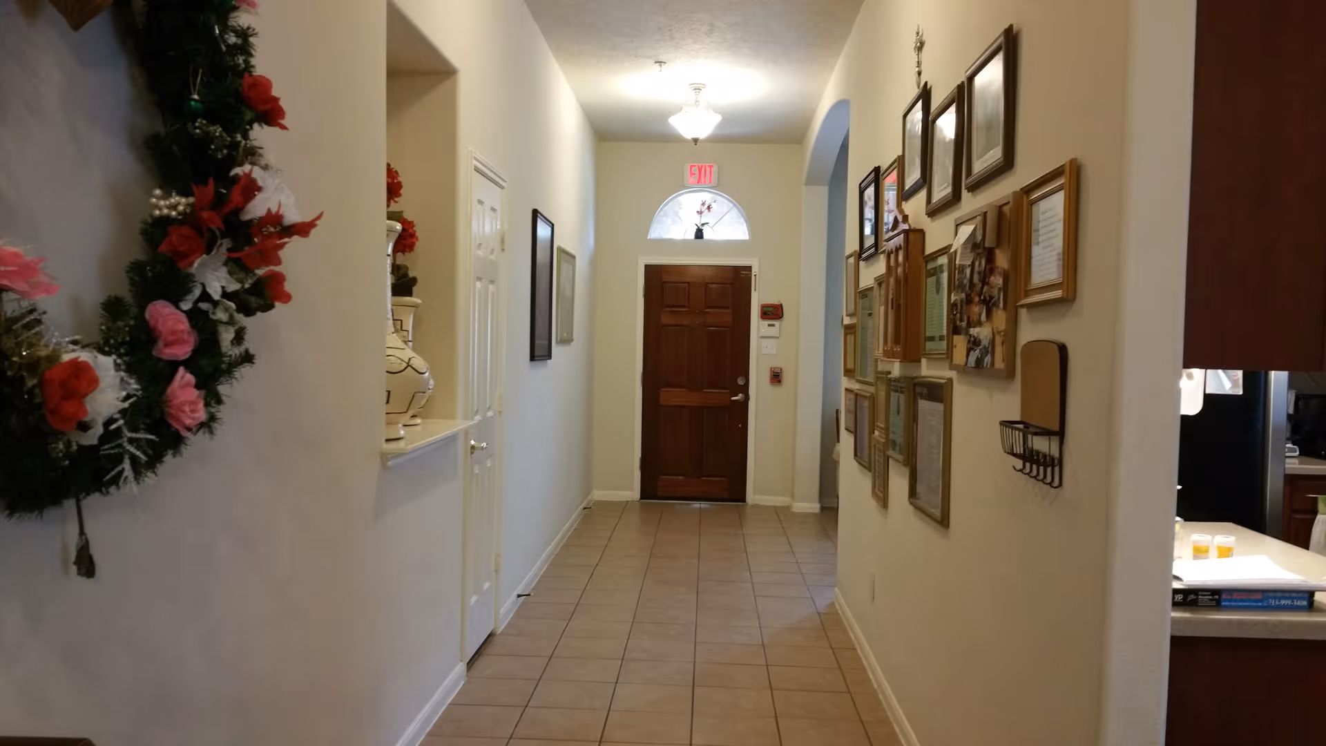 A hallway with beige tiled floor and cream-colored walls. On the left wall, there is a decorative wreath with red, pink, and white flowers. On the right wall, there are multiple framed pictures and documents. At the end of the hallway, there is a wooden door with a small arched window above it, and an exit sign above the door. To the right, part of a kitchen counter with some items on it is visible.