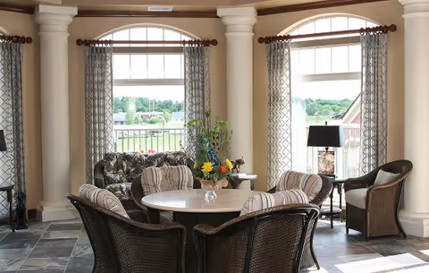 Bright sunroom-style sitting area with a round table surrounded by wicker chairs, sofas, decorative columns, and arched windows.