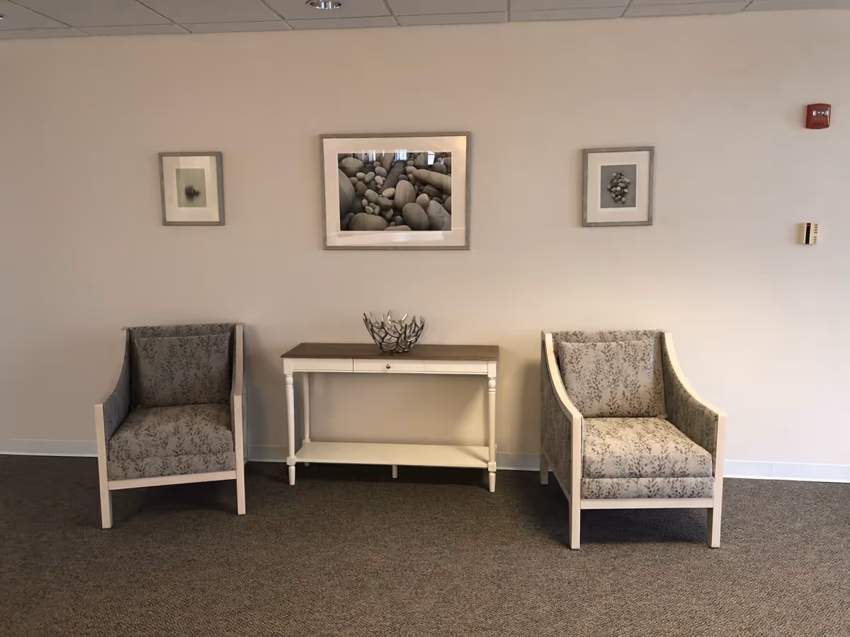 Two patterned armchairs with light wood frames are placed on either side of a white console table with a wooden top. On the table is a decorative metal bowl. Above the table, three framed pictures of stones and pebbles are hung on a plain beige wall. The floor is carpeted in a neutral tone.
