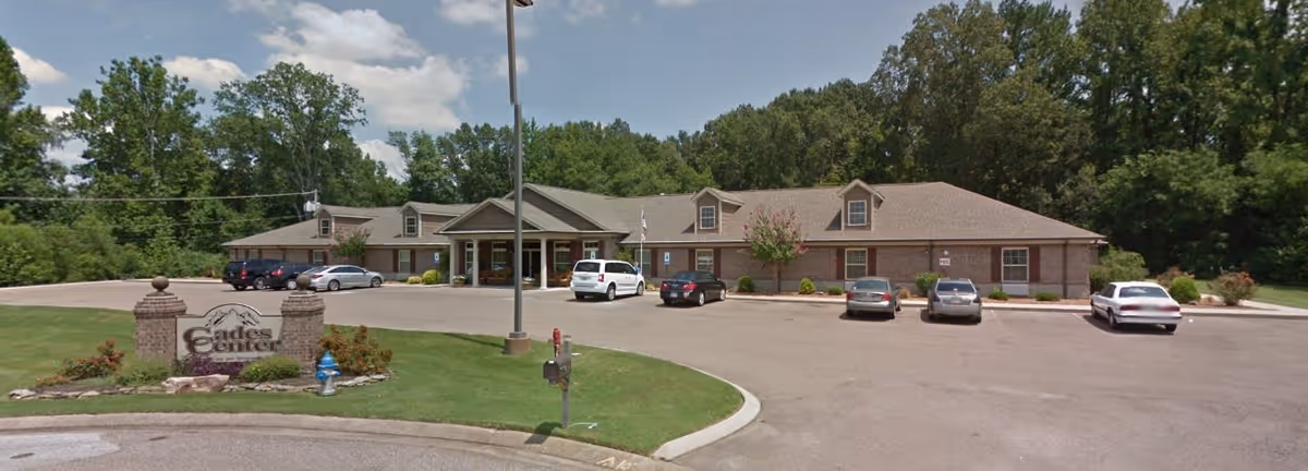 Exterior view of Cades Center Assisted Living, a single-story brick building with a parking lot in front. Several cars are parked, and there is a sign with the facility's name on a grassy area near the entrance. Trees and greenery surround the building under a partly cloudy sky.