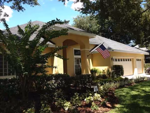 Yellow single-story senior living facility exterior with landscaped front yard and American flag.
