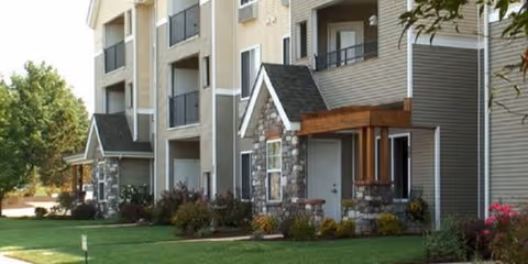 Exterior view of a multi-story senior living facility with stone and siding facade, small covered entryways, balconies, and well-maintained landscaping including grass, bushes, and trees.