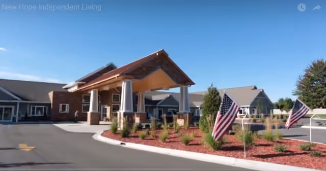 Front entrance of a senior living facility with a covered porte-cochere, landscaped beds with American flags, and surrounding single-story buildings under a blue sky.