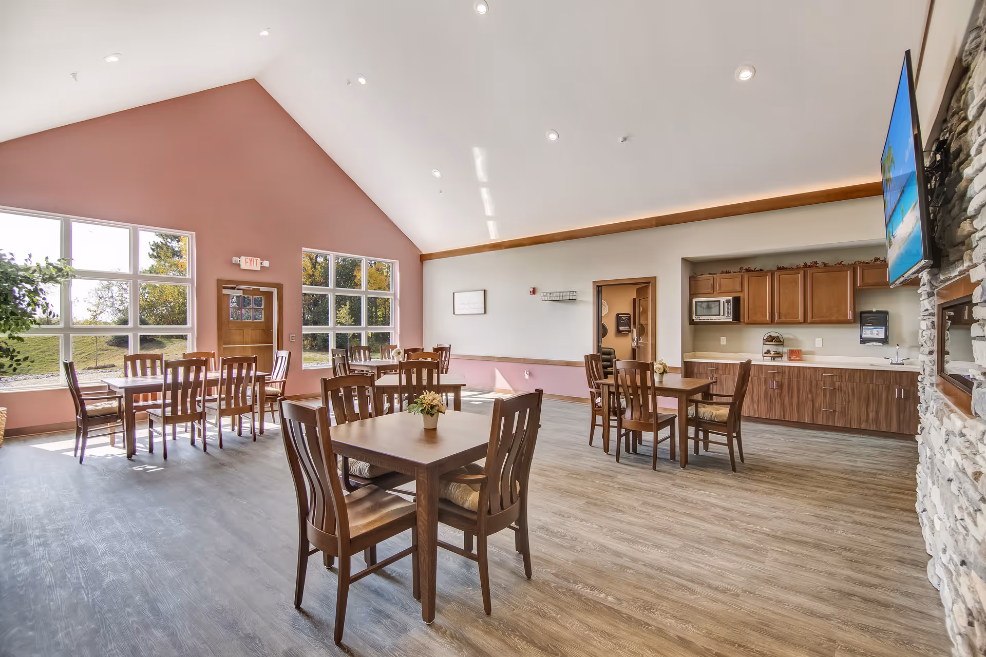 A bright and spacious dining room with several wooden tables and chairs arranged neatly. Large windows on one side allow natural light to fill the room, showing a view of greenery outside. The room features a high vaulted ceiling with recessed lighting, a small kitchenette area with cabinets, a microwave, and a sink. A flat-screen TV is mounted on a stone wall on the right side.