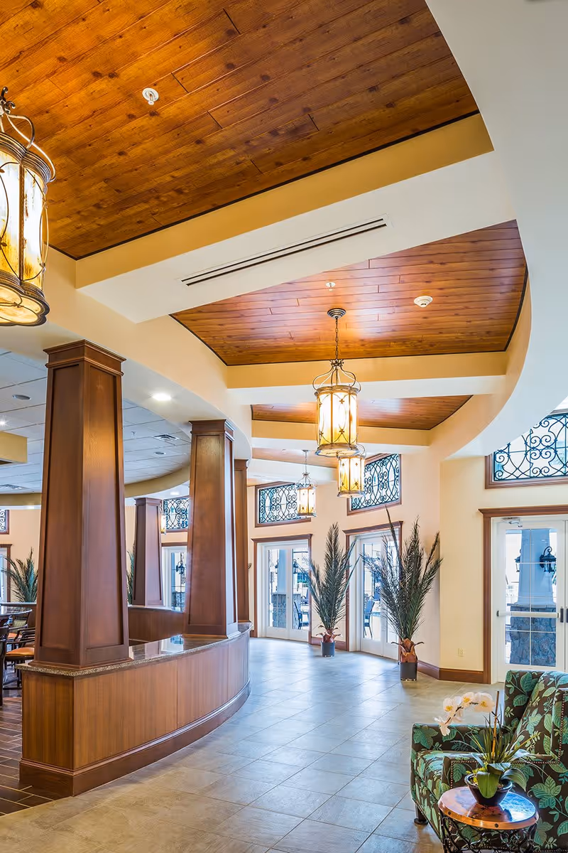 Bright and spacious interior of a senior living facility with wooden ceiling panels, hanging lantern-style lights, large windows with decorative ironwork, potted plants, and a curved wooden reception desk. There is a floral-patterned armchair and a small round table with a flower arrangement in the foreground.