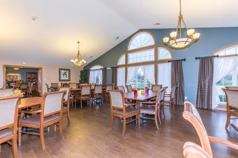 A spacious dining room with multiple wooden tables and chairs arranged neatly. The room features large arched windows with white curtains and dark drapes, allowing natural light to fill the space. Two chandeliers hang from the ceiling, illuminating the room. The floor is wooden, and there is a doorway leading to another room in the background.