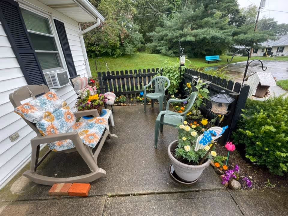 A small outdoor patio area with a black wooden fence, featuring two green plastic chairs, a beige rocking chair with floral cushions, and a small white side table. There are various potted plants and flowers around the patio, along with a bird feeder hanging on a metal hook. In the background, there is a grassy area with trees and a blue bench near a paved road.