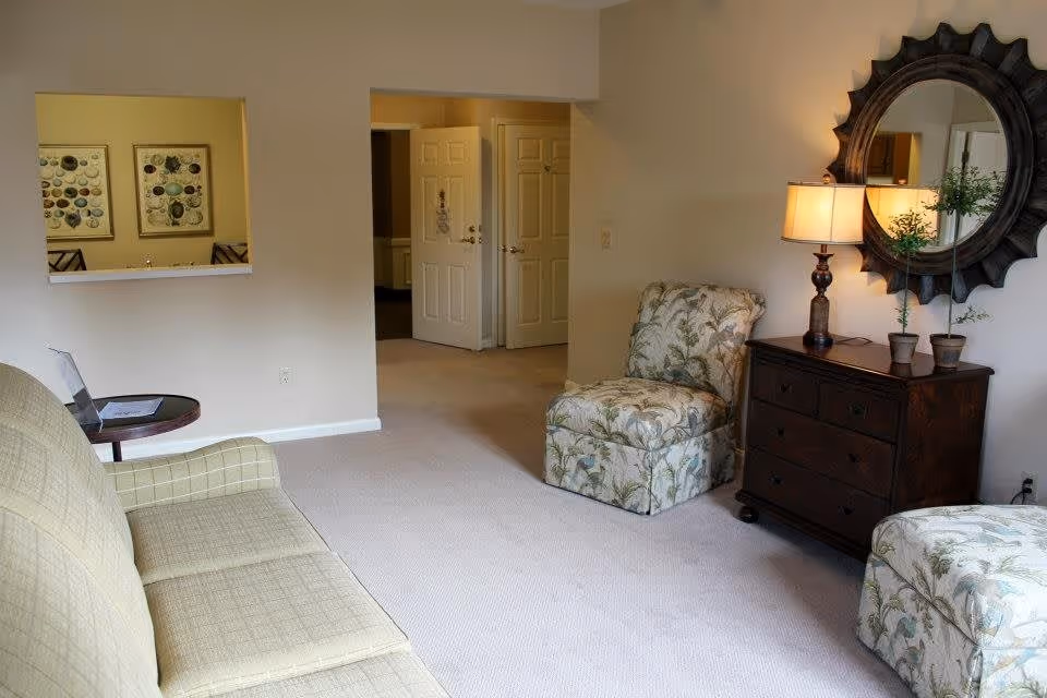 A cozy living room with a beige sofa on the left, a small round side table with a laptop and papers, two floral patterned chairs, a dark wooden dresser with a lamp and two small potted plants, and a large decorative round mirror on the wall. There is a doorway leading to another room and a pass-through window showing framed artwork in the adjacent room.
