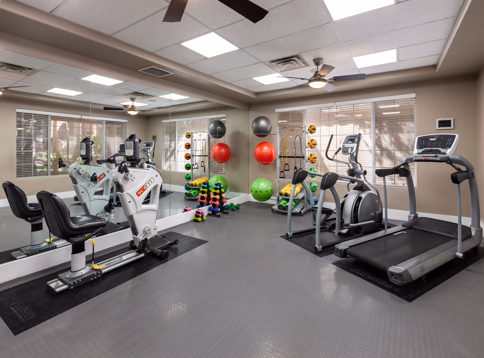 A well-lit fitness room with exercise equipment including a recumbent bike, elliptical machine, and treadmill. The room has large windows with blinds, a wall mirror, ceiling fans, and various exercise balls, dumbbells, and foam rollers neatly organized on racks.