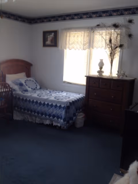 A small bedroom with a single bed covered in a blue and white quilt. There is a wooden headboard and a wooden chair next to the bed. A window with lace curtains lets in natural light. Next to the window is a wooden dresser with a decorative lamp and a vase with peacock feathers on top. The walls are white with a floral border near the ceiling.
