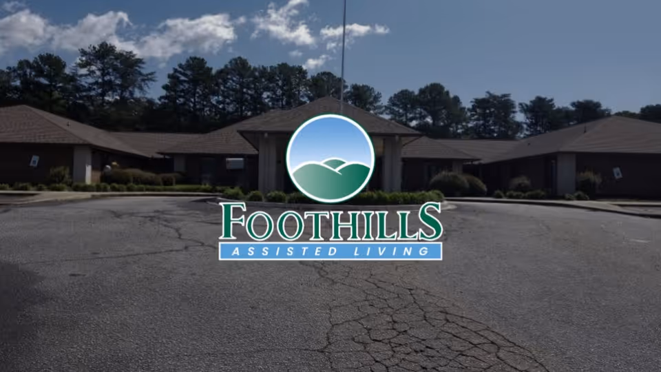 Exterior view of a single-story assisted living facility building with a circular driveway and trees in the background under a partly cloudy sky.