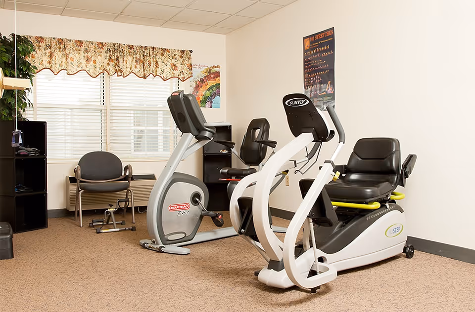 A small exercise room with two stationary exercise bikes, a chair, a small black shelving unit, and a window with floral valance curtains. There are two posters on the walls, one showing stretches and the other with a colorful design.