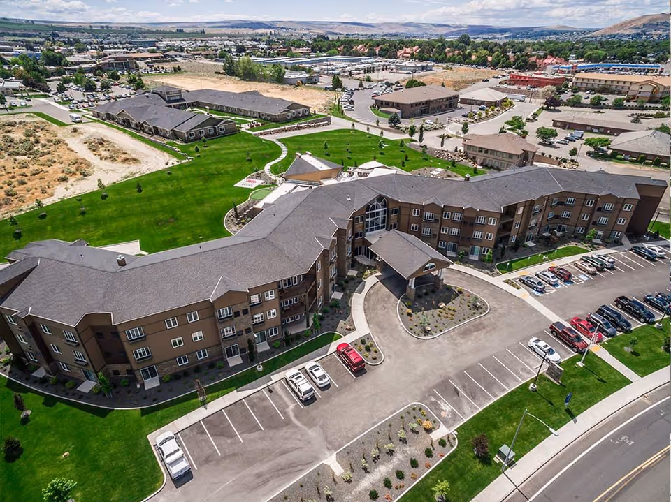 Aerial view of a senior living facility named Ciel of Tri-Cities showing multiple connected buildings with brown roofs and beige walls, surrounded by green lawns, parking lots with cars, and a curved driveway entrance. The background includes other buildings and a distant landscape with hills under a partly cloudy sky.