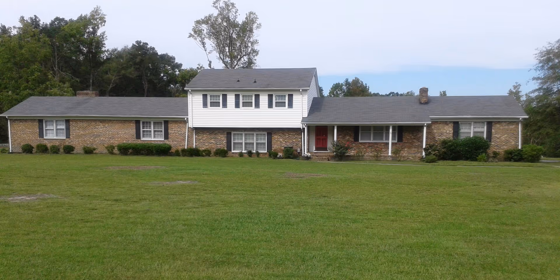 A single-story brick building with a central two-story white section, black shutters, and a red front door, surrounded by a large grassy lawn and trees in the background under a partly cloudy sky.