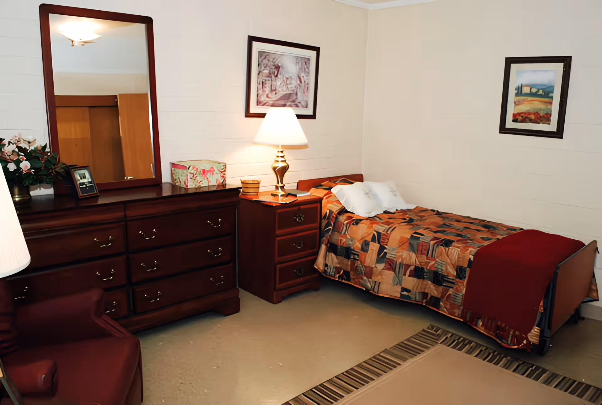 A small bedroom with a single bed covered in a patterned quilt and a red blanket at the foot. Next to the bed is a wooden nightstand with a lamp and some small items. On the left side of the room, there is a large wooden dresser with a mirror, a floral arrangement, a framed photo, and a decorative box on top. Two framed pictures hang on the beige walls. A maroon armchair is partially visible in the foreground.
