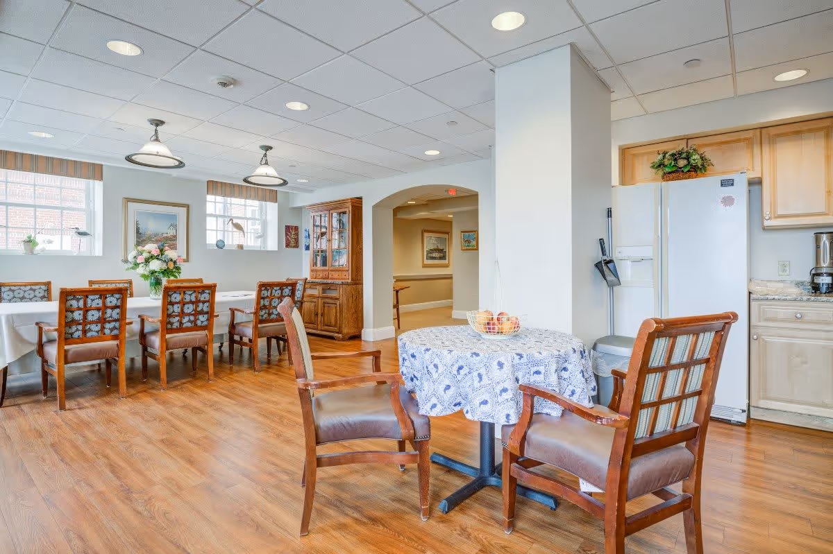 A bright dining area in a senior living facility featuring a long table with eight wooden chairs, a smaller round table with two chairs covered with a patterned tablecloth, a white refrigerator, wooden cabinets, and decorative elements including flowers and framed pictures on the walls.