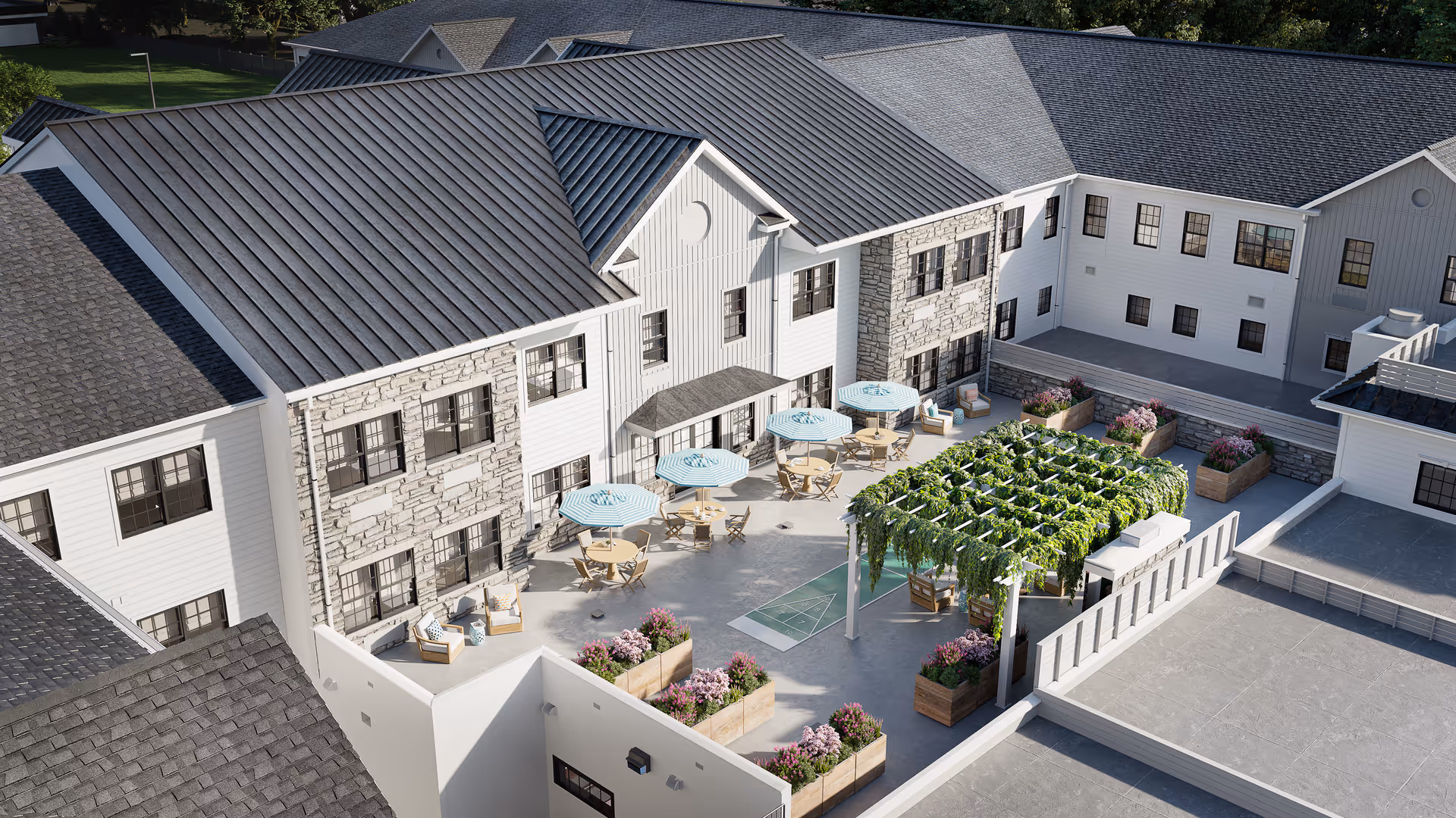 Aerial view of an outdoor courtyard at Azalea at Cinnaminson featuring multiple seating areas with tables and umbrellas, a pergola covered with greenery, planter boxes with flowers, and a shuffleboard court, surrounded by a two-story building with stone and white siding exterior.