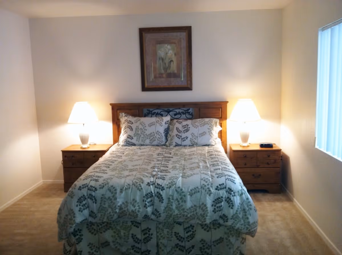 A bedroom with a bed centered against a wall, featuring a wooden headboard and a comforter with a leaf pattern. There are two matching pillows, two wooden nightstands on either side of the bed, each with a lit table lamp. A framed floral artwork hangs above the bed, and a window with vertical blinds is visible on the right side.