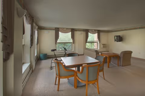 Common room with two tables and wooden chairs, lounge chairs, an exercise bike, and a wall-mounted TV beneath three windows.