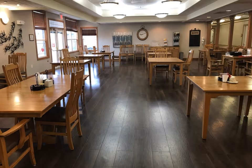 A spacious dining room with multiple wooden tables and chairs arranged neatly. The room has large windows with wooden blinds allowing natural light to enter. The walls are decorated with a metal bird sculpture, a large clock, and a painting. There is a beverage station and a chalkboard on the far wall. The floor is dark wood, and the ceiling has several round light fixtures.