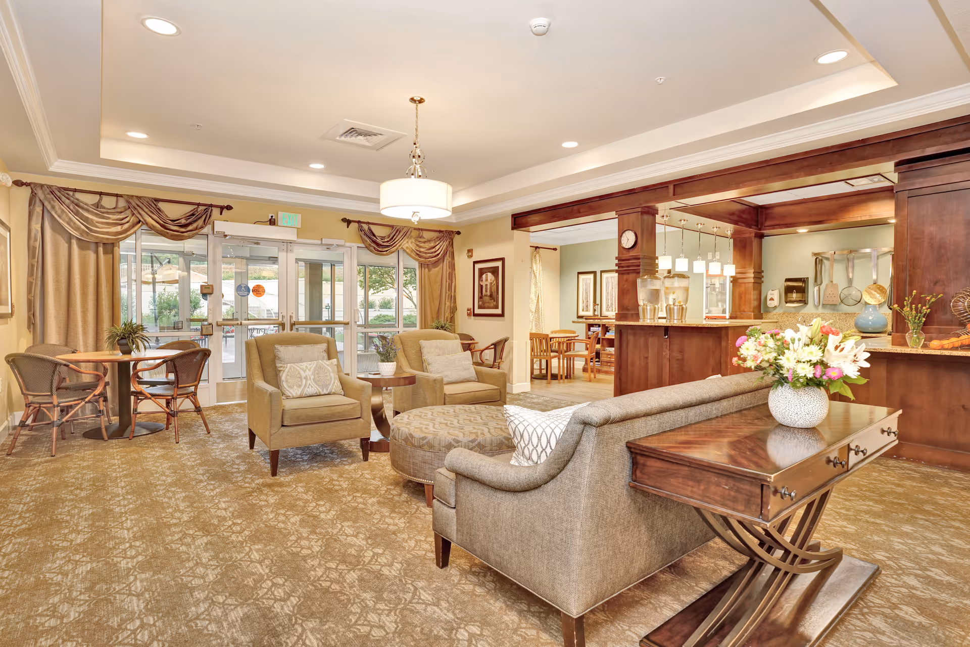 A spacious and well-lit common area in a senior living facility featuring comfortable armchairs, a sofa, a round ottoman, and a wooden side table with a vase of flowers. The room has large windows with draped curtains, a chandelier, and a view into a dining area with wooden tables and chairs. The decor includes warm tones, carpeted flooring, and wooden accents.