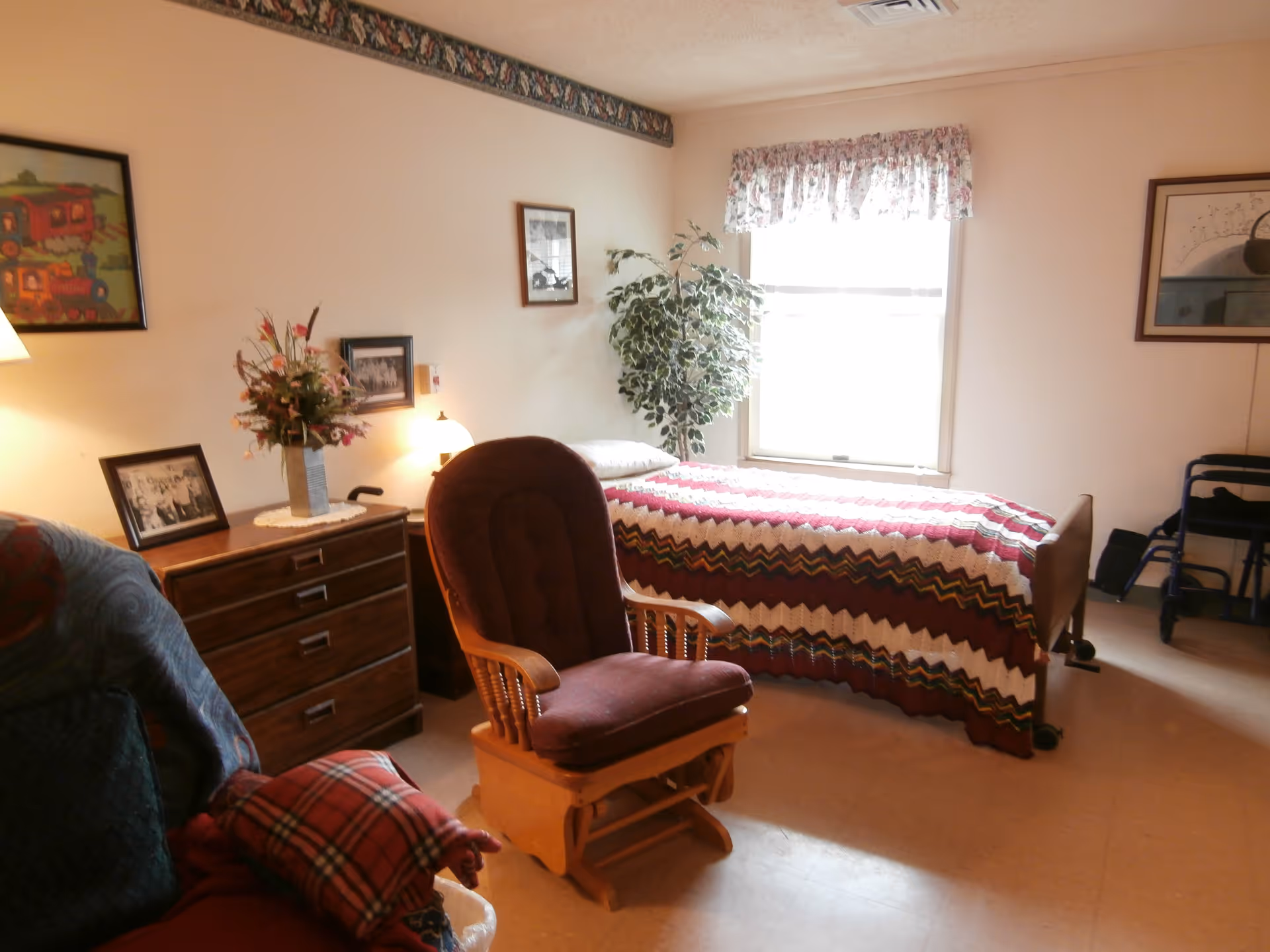 A cozy bedroom with a single bed covered by a colorful zigzag patterned blanket. There is a wooden rocking chair with a cushioned seat in front of the bed. A wooden dresser with a vase of flowers and framed pictures sits against the wall. A window with floral curtains lets in natural light. A walker is visible in the corner of the room.
