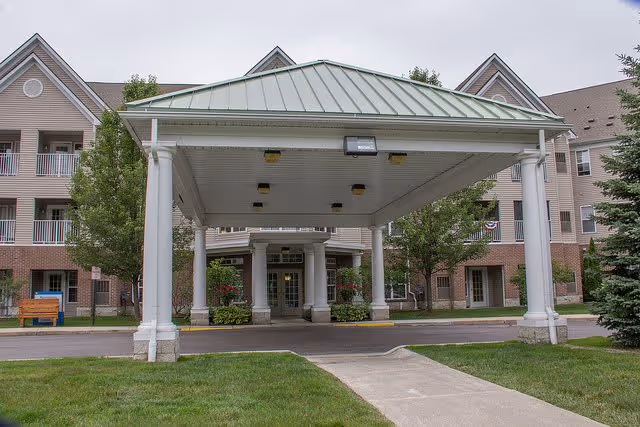 Entrance of a senior living facility with a covered driveway supported by white columns, surrounded by green grass, trees, and a multi-story building with balconies in the background.