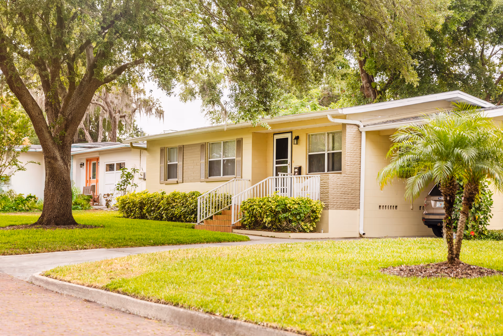 Front exterior of a single-story yellow house with a small porch, ramp, lawn, and large trees.
