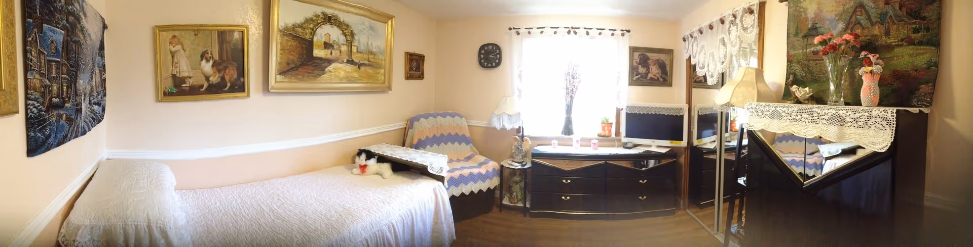 A cozy bedroom with a single bed covered in a white quilt, a stuffed cat toy on the bed, a chair with a crocheted blanket, a dresser with a TV, and various framed paintings and lace curtains on the window. The room has warm lighting and wooden flooring.