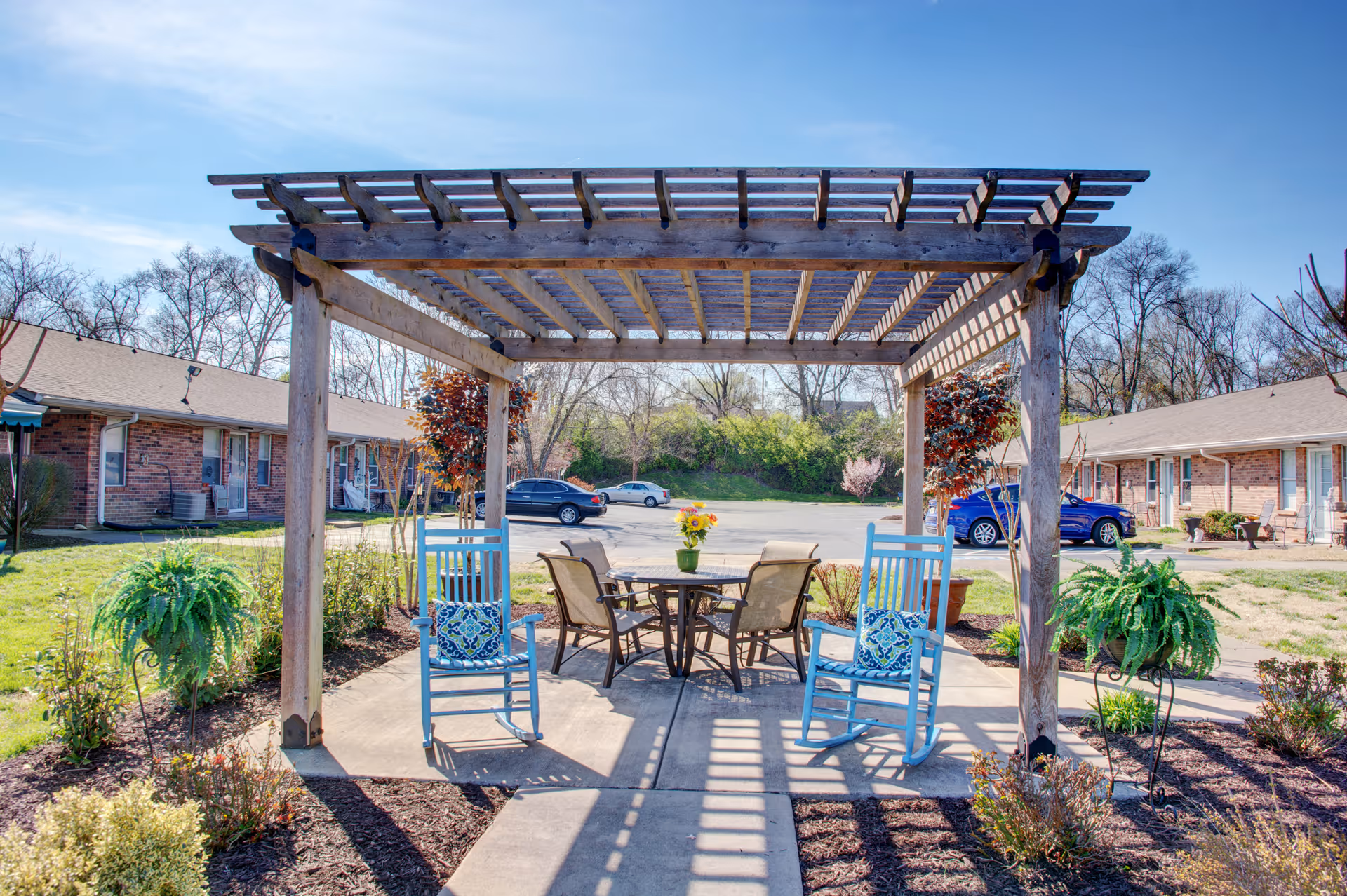 Outdoor seating area at Red Cedar Glen featuring a wooden pergola with two blue rocking chairs and a round table with four chairs. The table has a small vase with sunflowers. Surrounding the seating area are plants and shrubs, with brick buildings and parked cars visible in the background under a clear blue sky.