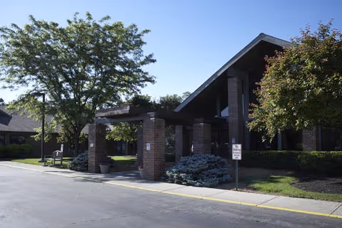 Exterior view of a senior living facility named Arbors at Milford showing a covered entrance with brick pillars, a driveway, trees, and landscaped bushes under a clear blue sky.