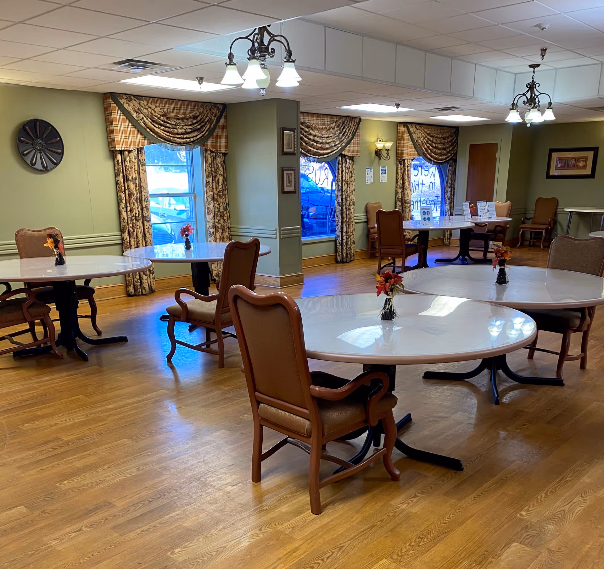 Bright dining room with several round tables, upholstered chairs, small floral centerpieces and windows with patterned curtains.