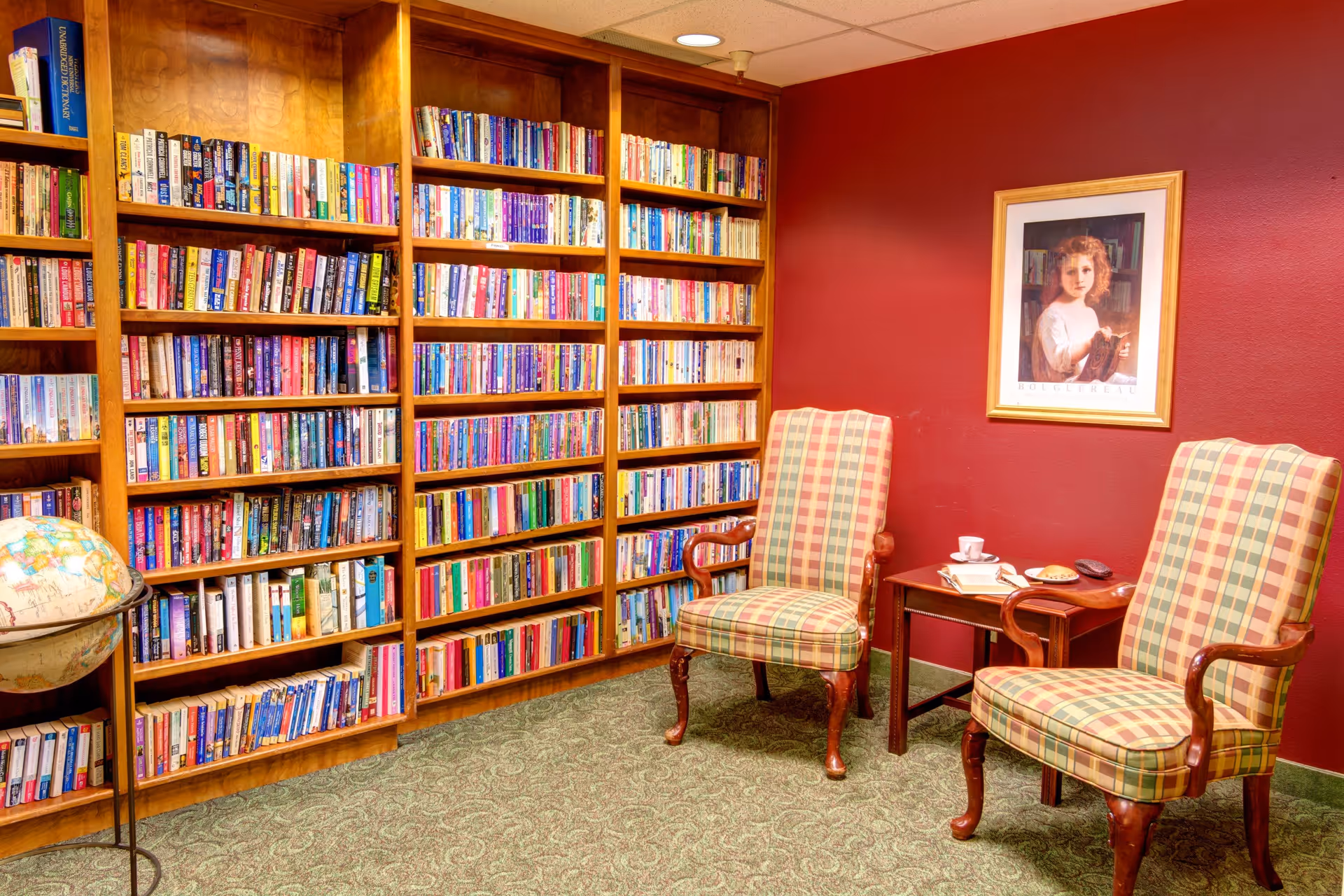 A cozy reading room with wooden bookshelves filled with books, two plaid upholstered armchairs, a small wooden side table with a cup, saucer, and a plate with a pastry, a globe on a stand, and a framed portrait of a young girl on a red wall.