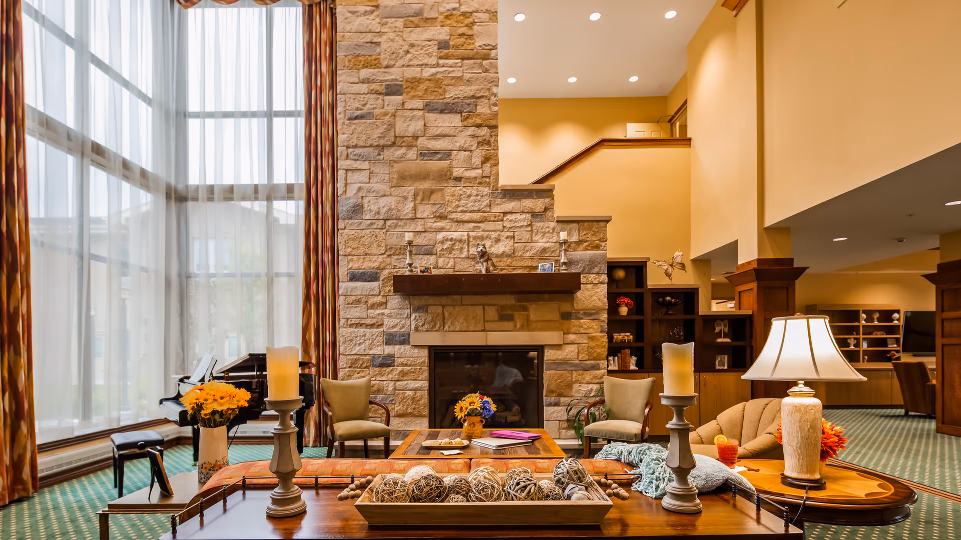 A spacious and warmly lit living room area with a large stone fireplace as the focal point. The room features tall windows with sheer curtains, a grand piano, comfortable armchairs, a sofa, a wooden coffee table decorated with candles and decorative balls, and a table lamp. The carpet is green with a subtle pattern, and the walls are painted in a soft beige tone.