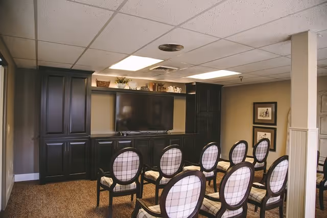 A small room with rows of chairs facing a large flat-screen TV mounted on a dark wooden cabinet. The cabinet has shelves with decorative items on top. The room has a beige carpet, beige walls, and a drop ceiling with fluorescent lights.