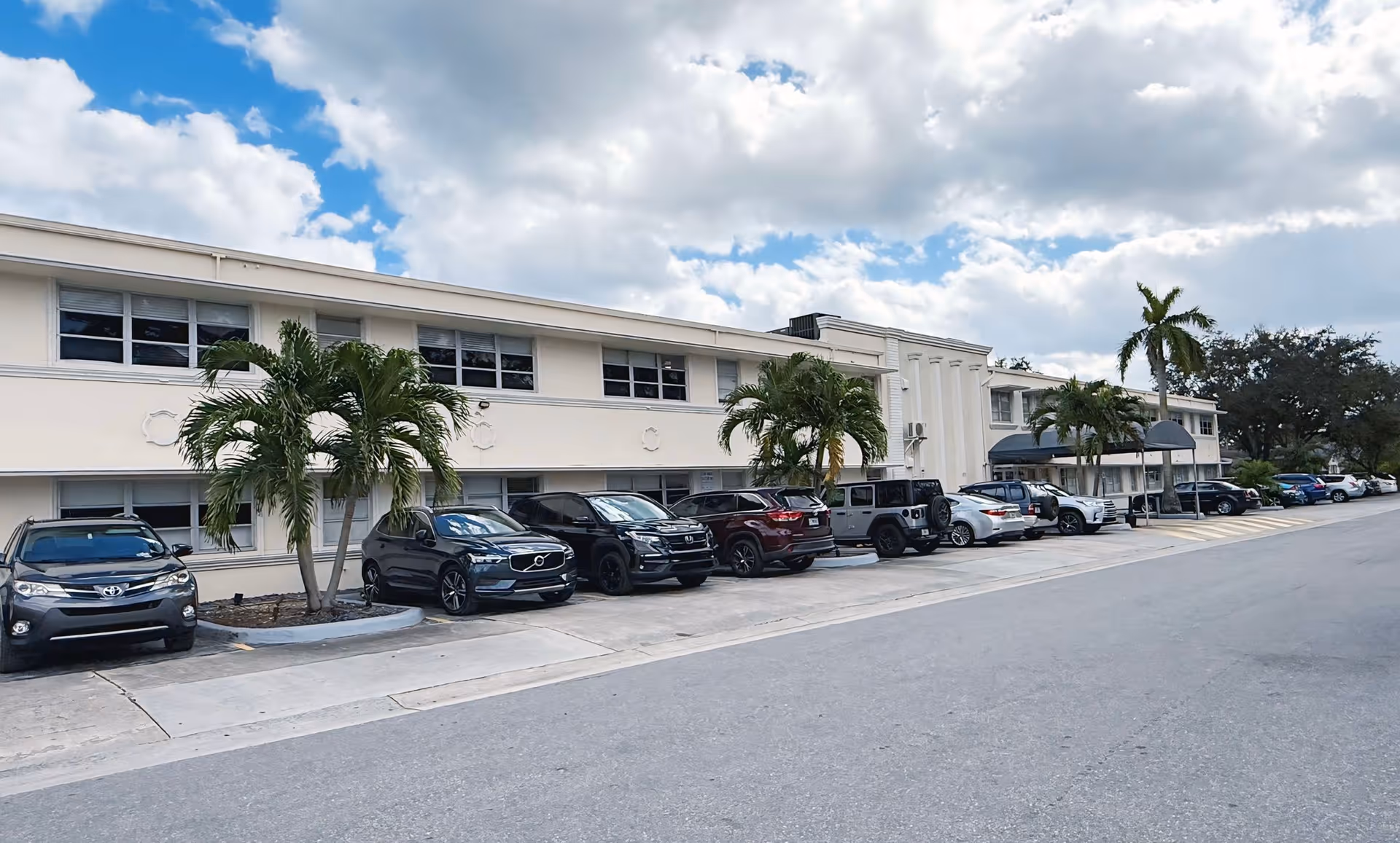 Exterior view of a two-story senior living facility building with several parked cars in front and palm trees along the sidewalk under a partly cloudy sky.