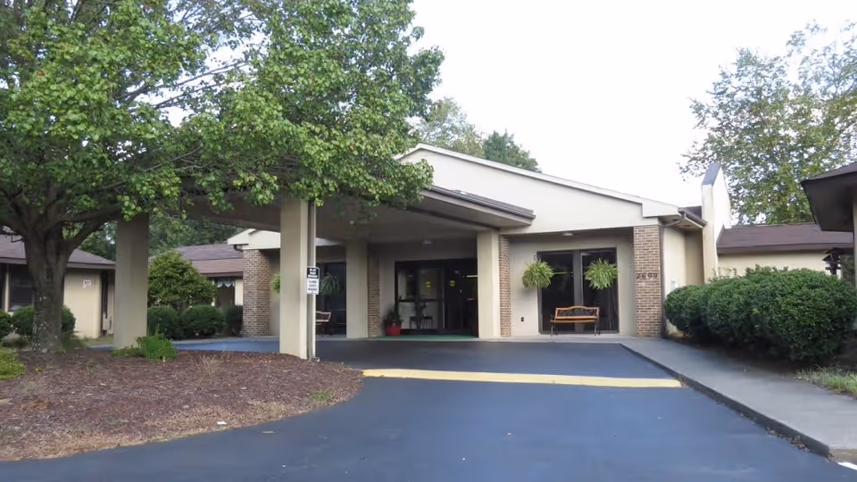 Front exterior view of Grand Villa Assisted Living at Winston showing the entrance with a covered drop-off area, surrounded by trees and bushes.