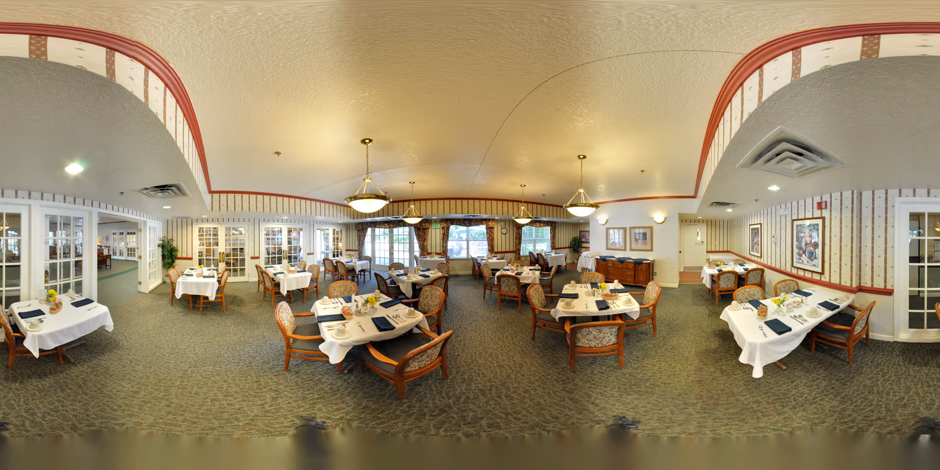 A spacious dining room in Oak Woods Manor with multiple tables set for meals. Each table is covered with a white tablecloth and has place settings including plates, napkins, and glasses. The room features patterned carpet, wallpaper with a red border near the ceiling, and several hanging light fixtures. Large windows with curtains allow natural light to enter, and there are framed pictures on the walls. The room has a warm and inviting atmosphere.