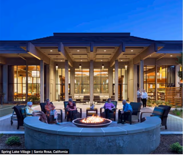 Outdoor patio area at dusk with a circular fire pit surrounded by six elderly people sitting on chairs. The background shows a modern building with large windows and warm interior lighting. Two people are walking near the entrance of the building.