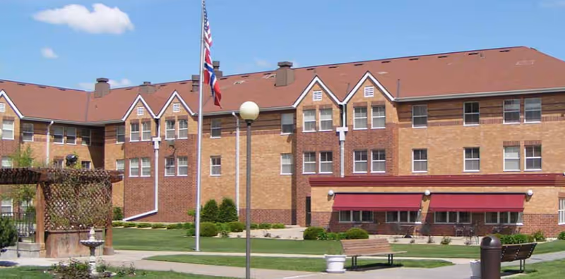 Exterior view of a large brick senior living facility building with multiple windows and a red roof. In front of the building is a well-maintained lawn with a flagpole displaying the American and another flag, benches, a lamp post, and a small pergola structure with a fountain nearby. The sky is clear with a few clouds.