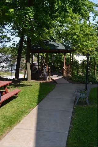 A shaded outdoor pathway leading to a wooden gazebo surrounded by green trees and grass. There is a red picnic table on the left side of the path and a metal bench on the right side. A lamp post is also visible near the bench.
