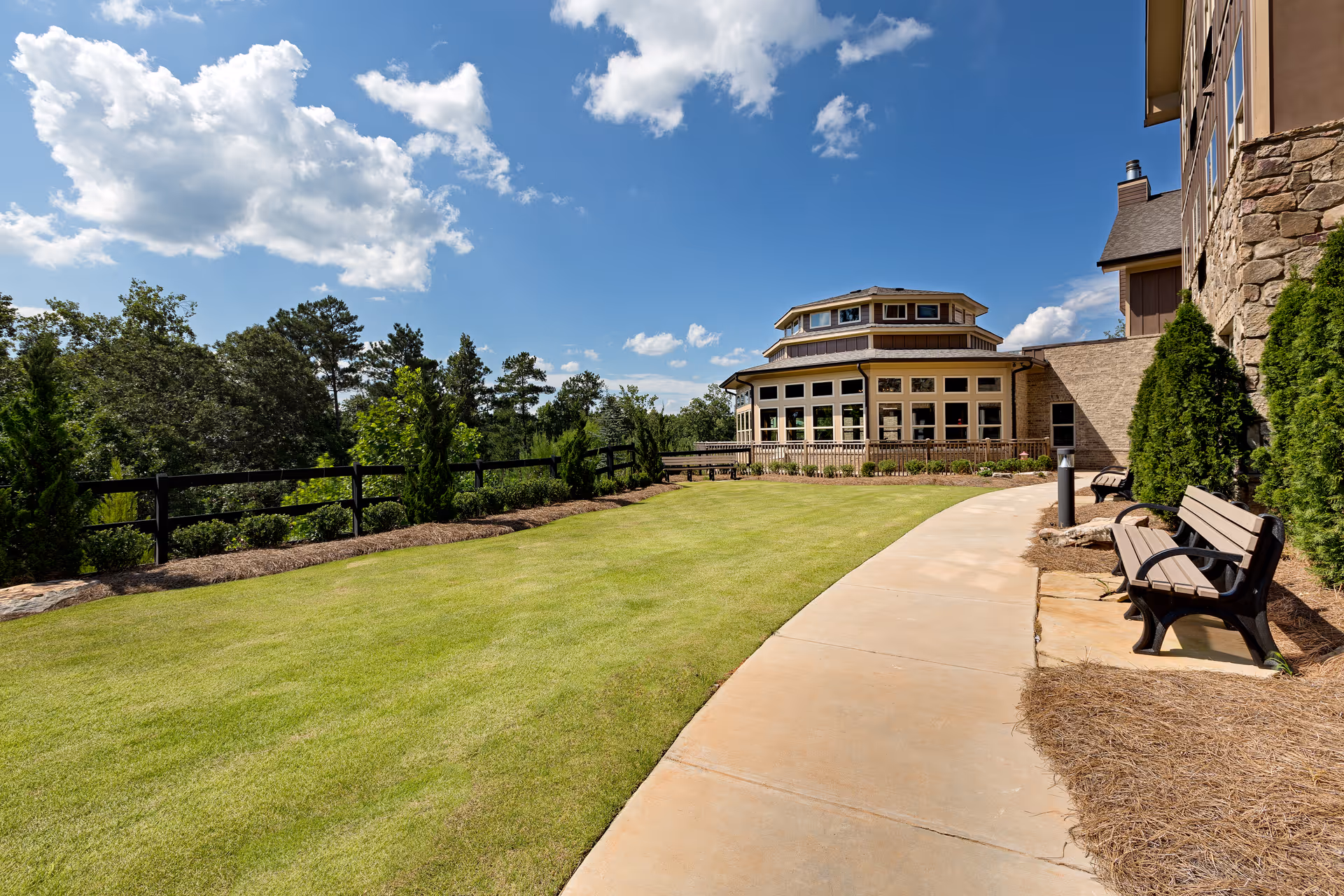 Outdoor area of Oaks at Acworth senior living facility featuring a curved concrete walkway, green lawn, benches along the path, and a building with large windows under a blue sky with scattered clouds.