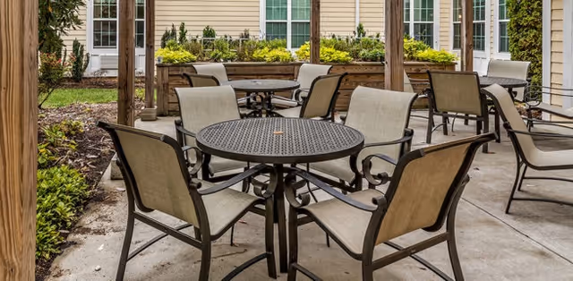 Outdoor patio area with multiple round metal tables and beige cushioned chairs under a wooden pergola, surrounded by greenery and the exterior walls of a building with windows and doors.