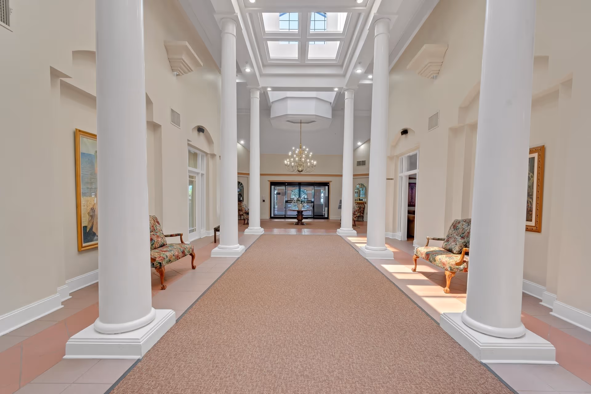 A spacious, well-lit hallway in a senior living facility with tall white columns on both sides, floral upholstered benches, framed artwork on the walls, a large chandelier hanging from the ceiling, and a skylight allowing natural light to brighten the area. The hallway leads to glass doors at the far end.