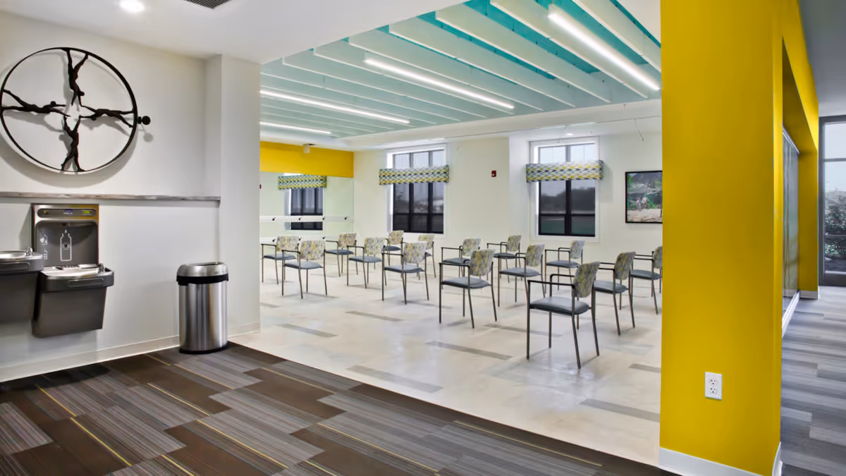 A spacious, well-lit room with multiple chairs arranged in rows, likely for meetings or group activities. The room features large windows with patterned valances, a yellow accent wall, and a modern ceiling with linear lighting. In the foreground, there are two water fountains and a trash can against a white wall with a decorative circular metal art piece.