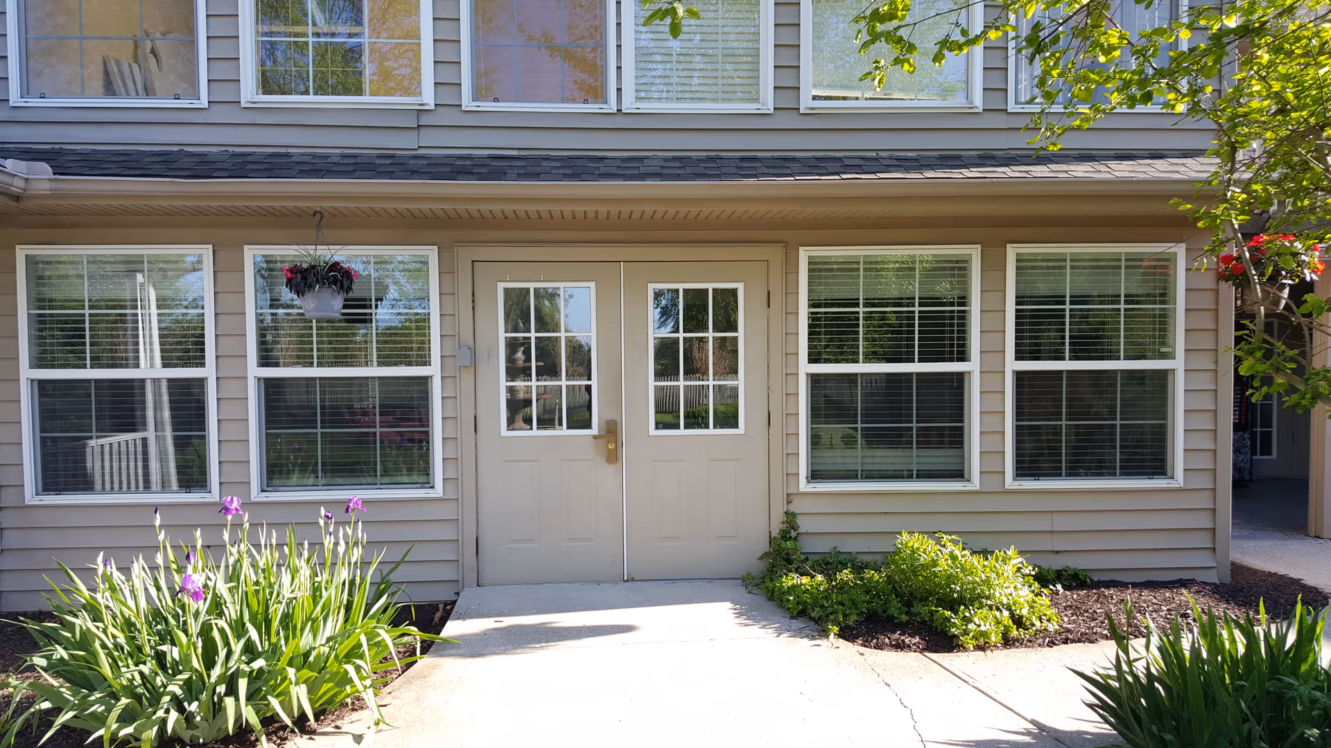 Front entrance of a beige building with double doors, large windows, and landscaped flowerbeds.